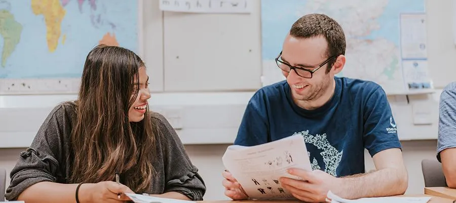 Image of two students learning a language at a desk, holding paper